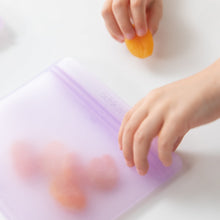 Load image into Gallery viewer, Close-up of a child's hands placing an apricot into a Bumkins Silicone Flat Reusable Bag 2 Pack, Lavender on a white table.