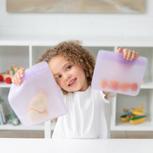 Load image into Gallery viewer, A curly-haired child happily holds Bumkins Silicone Flat Reusable Bag 2 Pack, Lavender full of snacks at a white table.