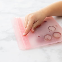 Load image into Gallery viewer, A hand presses on a Bumkins Silicone Flat Reusable Bag 2 Pack, Pink filled with fruit on a marble surface, sealing the leak-proof closure.