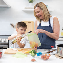 Load image into Gallery viewer, A woman holds a Bumkins Silicone Flat Reusable Bag 2 Pack, Sage with salmon as a boy pours oil, surrounded by fresh ingredients in a kitchen.