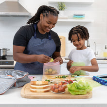 Load image into Gallery viewer, A woman and child prepare lunch using Bumkins Silicone Flat Reusable Bag 2 Pack, Sage at a kitchen counter surrounded by fresh ingredients.