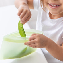 Load image into Gallery viewer, A child smiling in a white shirt, placing peas into a Bumkins Silicone Flat Reusable Bag 2 Pack, Sage at a white table.