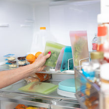 Load image into Gallery viewer, A hand places a Bumkins Silicone Flat Reusable Bag 2 Pack, Sage filled with veggies in a fridge, with other bags, a milk jug & fruit visible.