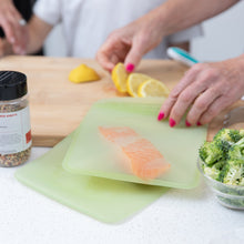 Load image into Gallery viewer, A person prepares a meal with salmon & lemon slices using Bumkins Silicone Flat Reusable Bag 2 Pack, Sage on a kitchen counter.