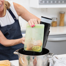 Load image into Gallery viewer, A person in an apron places a sealed Bumkins Silicone Flat Reusable Bag 2 Pack, Sage with salmon and lemon into a pot with a sous vide device.