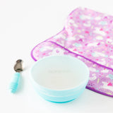 Bumkins Silicone Grip Bowl: Blue and a blue spoon sit in front of a purple unicorn bib on a white background, covered with a silicone lid.