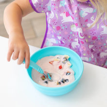 Load image into Gallery viewer, A child eats yogurt and fruit from Bumkins Silicone Grip Bowl: Blue, wearing a purple unicorn bib at a white table.