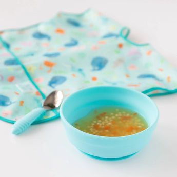Bumkins Silicone Grip Bowl: Blue with soup and pasta sits in front of a spoon and a blue bib on a white background.