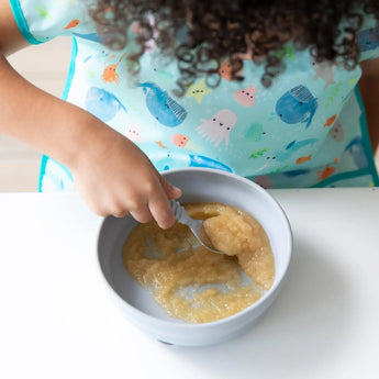 A child enjoys applesauce from Bumkins Silicone Grip Bowl: Gray, spoon in hand, wearing a colorful ocean-themed bib at a white table.