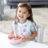 A smiling toddler with brown hair in a floral bib enjoys cereal from a Bumkins Silicone Grip Bowl: Pink with a pink spoon at a white table.