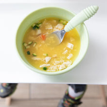 Load image into Gallery viewer, Bumkins Silicone Grip Bowl: Sage with chicken soup and a green spoon sits on a high chair tray; a child's feet are visible below.