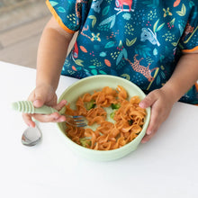 Load image into Gallery viewer, A child in a colorful bib eats pasta from a Bumkins Silicone Grip Bowl: Sage with a fork at a white table.
