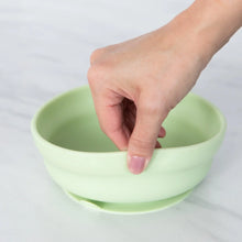 Load image into Gallery viewer, A hand holds a Bumkins Silicone Grip Bowl: Sage, demonstrating the suction cup bottom on a marble surface.