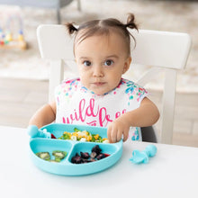Load image into Gallery viewer, A toddler with pigtails enjoys a meal from a Bumkins Silicone Grip Dish: Blue, with sections of fruits and veggies, at a white table.