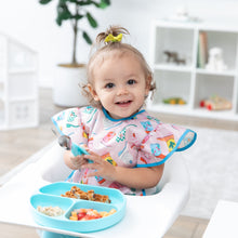 Load image into Gallery viewer, A girl in a high chair smiles, holding Bumkins Spoon + Fork: Blue by a blue divided plate of food with toy-filled shelves behind.
