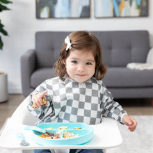 Load image into Gallery viewer, A girl wearing a Bumkins Sleeved Bib: Charcoal Check smirks in a high chair, eating a meal from a blue plate with matching utensils.