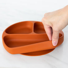 Load image into Gallery viewer, A hand holds a Bumkins Silicone Grip Dish: Clay, demonstrating the suction cup bottom on a marble surface.
