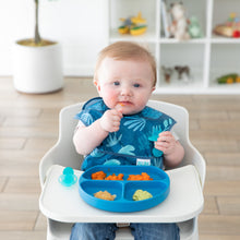Load image into Gallery viewer, A baby in a blue bib sits in a high chair, practicing self-feeding with Bumkins Silicone Grip Dish: Dark Blue in front of toy-filled shelves.