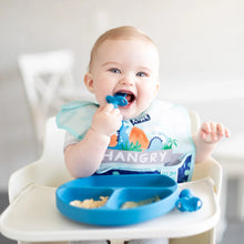 Load image into Gallery viewer, A smiling baby in a high chair wears a dinosaur bib, holding a spoon while eating from a Bumkins Silicone Grip Dish: Dark Blue.