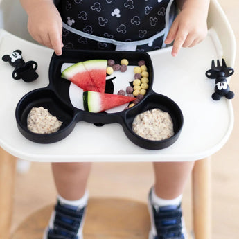 Close-up of a child in a high chair with cereal, fruit, and oatmeal in a Bumkins Silicone Grip Dish: Mickey Mouse; utensils sit close by.