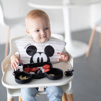 A toddler in a high chair enjoys strawberries and bananas from a Bumkins Silicone Grip Dish: Mickey Mouse while wearing a matching bib.