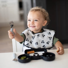 Load image into Gallery viewer, A curly-haired toddler with a Bumkins Silicone Grip Dish: Mickey Mouse holds a fork at a white table, wearing a matching bib.