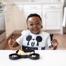 Load image into Gallery viewer, A toddler at a white table in a kitchen eagerly holds utensils over a Bumkins Silicone Grip Dish: Mickey Mouse with food.