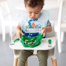 Load image into Gallery viewer, A toddler in a high chair wearing a dinosaur bib lifts a Bumkins Silicone Grip Dish, Special Edition: Dino with food attached to the tray.