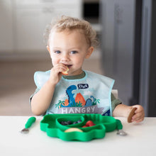 Load image into Gallery viewer, A curly-haired child smiles as they eat from Bumkins Silicone Grip Dish, Special Edition: Dino, wearing a dinosaur bib at a white table.