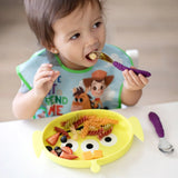 A toddler self-feeds pasta and fruit from a Bumkins Silicone Grip Dish: Toy Story Alien, wearing a bib with cartoon characters.