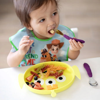 A toddler self-feeds pasta and fruit from a Bumkins Silicone Grip Dish: Toy Story Alien, wearing a bib with cartoon characters.
