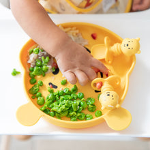 Load image into Gallery viewer, Close-up of a child's hand reaching for peas from a Bumkins Silicone Grip Dish: Winnie The Pooh, with matching utensils on top.