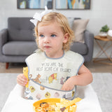 A girl in a bow in a high chair holds a Bumkins Spoon + Fork: Winnie the Pooh by a plate of breakfast food,reaching into her bib pocket.