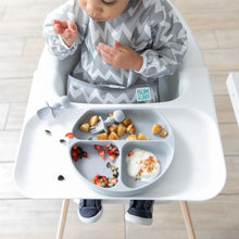 Load image into Gallery viewer, A baby in a high chair wearing a gray zig-zag bib enjoys food from a Bumkins Silicone Grip Dish: Gray with matching utensils.