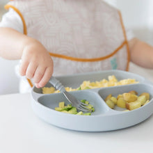 Load image into Gallery viewer, Close-up of a child in a beige bib enjoying veggies with a fork from a Bumkins Silicone Grip Dish: Gray at a white table.