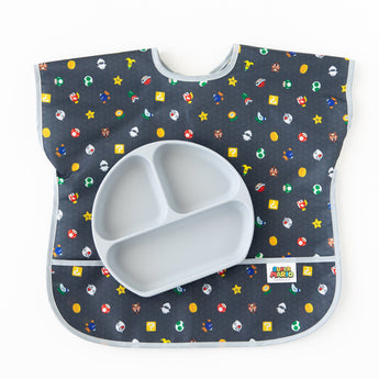 A gray divided plate sits on top of a Bumkins Junior Bib: Super Mario™ Lineup on a white background.
