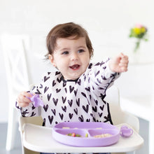 Load image into Gallery viewer, A smiling toddler in a high chair, wearing a heart-print bib, reaches toward the camera with a Bumkins Silicone Grip Dish: Lavender nearby.