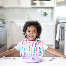 Load image into Gallery viewer, A toddler smiles in a bright kitchen, wearing a colorful bib at a white table with a Bumkins Silicone Grip Dish: Lavender and utensils.