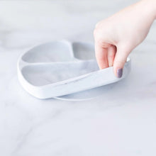 Load image into Gallery viewer, A hand holds a Bumkins Silicone Grip Dish: Marble, demonstrating the suction cup bottom on a marble surface.