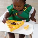 A baby in a high chair enjoys a meal from a Bumkins Silicone Grip Dish: Zelda™ Triforce with yellow utensils.