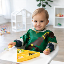 Load image into Gallery viewer, A toddler in a green bib sits in a high chair, gripping a fork and spoon next to a Bumkins Silicone Grip Dish: Zelda™ Triforce on the tray.