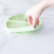 Load image into Gallery viewer, A hand holds a Bumkins Silicone Grip Dish: Sage, demonstrating the suction cup bottom on a marble surface.