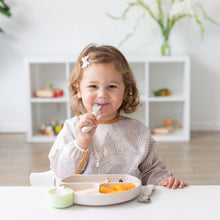 Load image into Gallery viewer, A toddler with a bow clip enjoys a meal from Bumkins Silicone Grip Dish: Sand, securely attached to the white table by its suction base.
