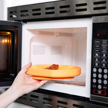 Load image into Gallery viewer, A person places a Bumkins Silicone Grip Dish: Tangerine with food into an open microwave.