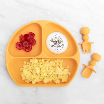 Bumkins Silicone Grip Dish: Tangerine holds berries, yogurt, and scrambled eggs next to orange toddler utensils on a marble background.