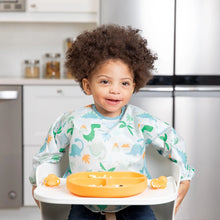 Load image into Gallery viewer, A curly-haired child in a dinosaur bib smiles in a high chair with a Bumkins Silicone Grip Dish: Tangerine and orange utensils on the tray.