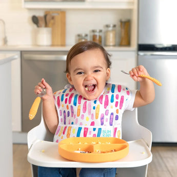 A toddler in high chair, wearing a colorful bib, smiles while holding orange utensils by a Bumkins Silicone Grip Dish: Tangerine.