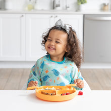 Load image into Gallery viewer, A smiling girl with a bow wearing a blue bib eats from a Bumkins Silicone Grip Dish: Tangerine with matching utensils in a kitchen.
