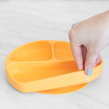 Load image into Gallery viewer, A hand holds a Bumkins Silicone Grip Dish: Tangerine, demonstrating the suction cup bottom on a marble surface.