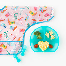 Load image into Gallery viewer, Bumkins Sleeved Bib: Boots lays flat next to a blue divided plate with veggies and rice and matching utensils on a white background.